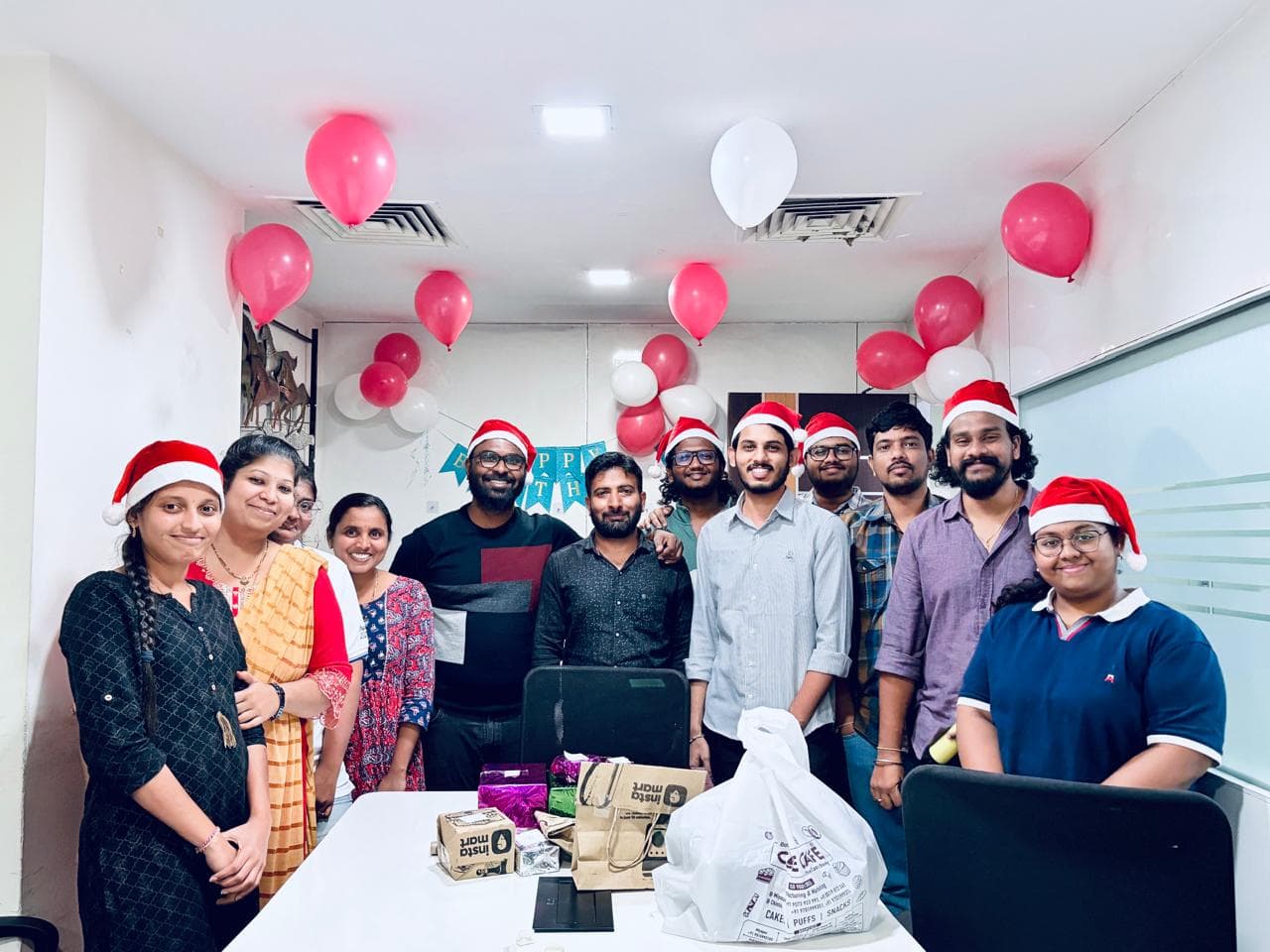 Colleagues wearing Santa hats at a Christmas office celebration with balloons and gifts