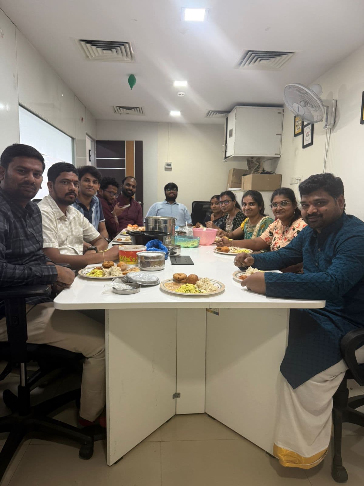 The team in traditional dress sharing a potluck lunch around the boardroom table, plates and tiffin carriers between them